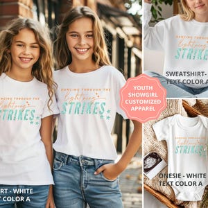 May include: White apparel including a t-shirt, a sweatshirt, and a onesie, each featuring the text Dancing Through The Lightning Strikes in orange and teal. The design includes small star accents. The items are displayed on individuals and in a basket.