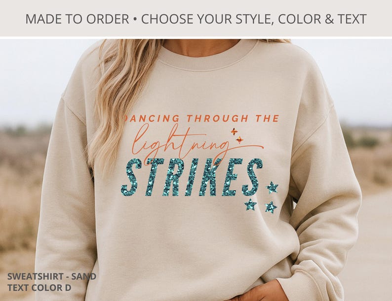 May include: A sand-colored crewneck sweatshirt featuring the text DANCING THROUGH THE lightning STRIKES in orange and teal glittery font. The design includes small orange and teal star accents. The top text reads MADE TO ORDER &bull; CHOOSE YOUR STYLE, COLOR & TEXT.