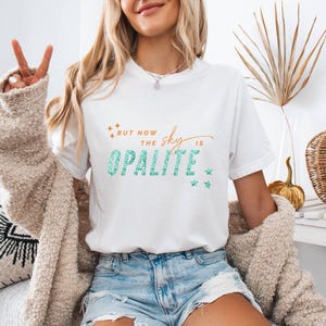 May include: White t-shirt with the text "But now the sky is Opalite" in teal and orange. The shirt has a crew neck and short sleeves. The model is wearing blue denim shorts and a beige cardigan.