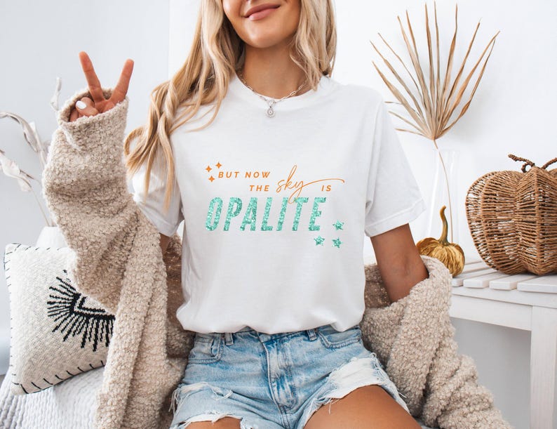 May include: White t-shirt with the text "But now the sky is Opalite" in teal and orange. The shirt has a crew neck and short sleeves. The model is wearing blue denim shorts and a beige cardigan.