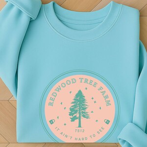 Pode incluir: Camisola azul clara com um design circular. O design inclui uma &aacute;rvore verde e as palavras "Redwood Tree Farm" em um c&iacute;rculo. O gr&aacute;fico est&aacute; em um fundo de p&ecirc;ssego com o texto "It ain't hard to see."