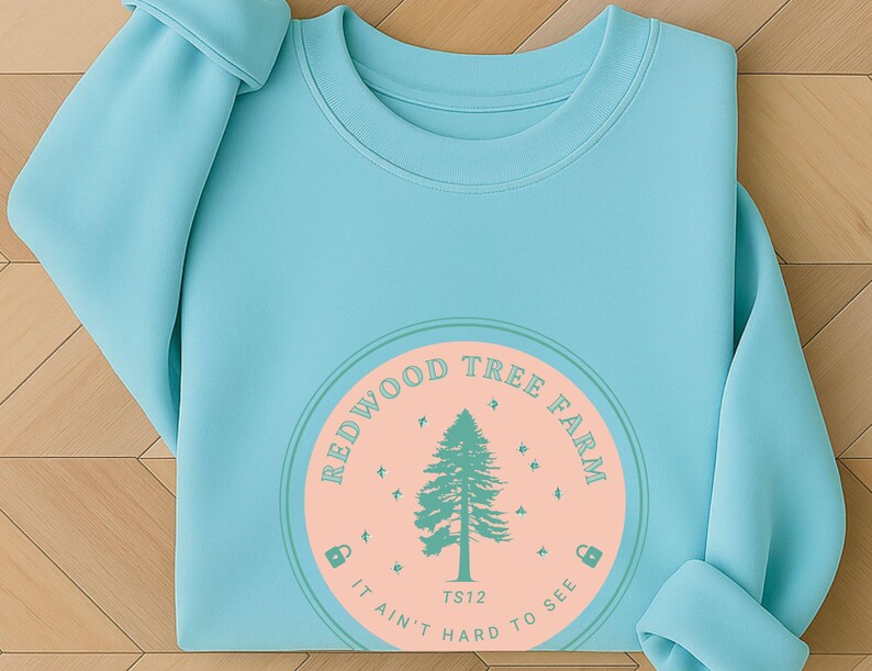 Pode incluir: Camisola azul clara com um design circular. O design inclui uma &aacute;rvore verde e as palavras "Redwood Tree Farm" em um c&iacute;rculo. O gr&aacute;fico est&aacute; em um fundo de p&ecirc;ssego com o texto "It ain't hard to see."