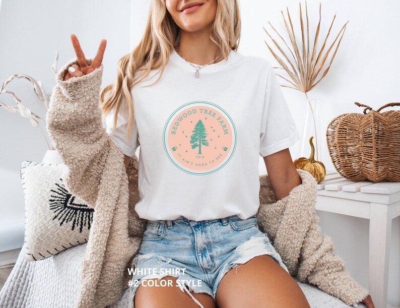 Pode incluir: Camiseta branca com um gr&aacute;fico circular com uma sequoia e o texto "Redwood Tree Farm 1912 It ain't hard to see". A camiseta &eacute; combinada com shorts jeans. A imagem tamb&eacute;m mostra um su&eacute;ter bege.