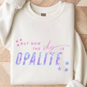 May include: Cream-colored sweatshirt with the text "But now the sky is Opalite" in a mix of purple and pink hues. The word "Opalite" has a geometric pattern. Pink and blue star graphics are also present.