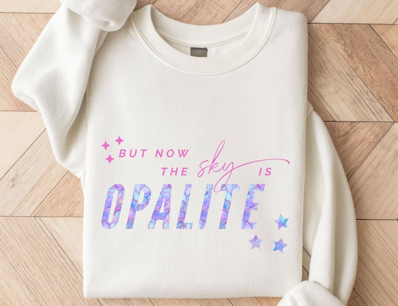 May include: Cream-colored sweatshirt with the text "But now the sky is Opalite" in a mix of purple and pink hues. The word "Opalite" has a geometric pattern. Pink and blue star graphics are also present.