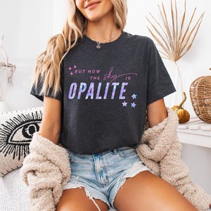 May include: Dark gray t-shirt with the text "But now the sky is Opalite" in a purple and blue gradient. The shirt features a crew neck and short sleeves. The design includes star graphics. The shirt is made of a soft material.