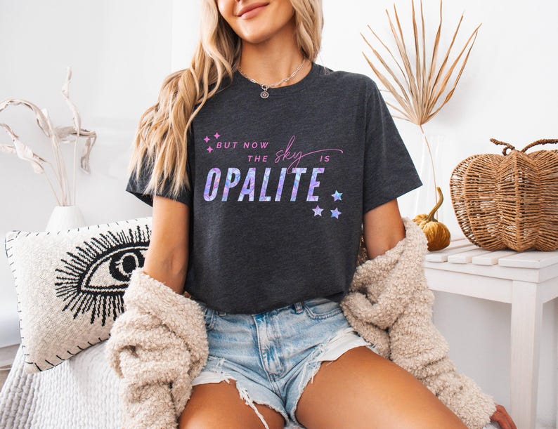 May include: Dark gray t-shirt with the text "But now the sky is Opalite" in a purple and blue gradient. The shirt features a crew neck and short sleeves. The design includes star graphics. The shirt is made of a soft material.
