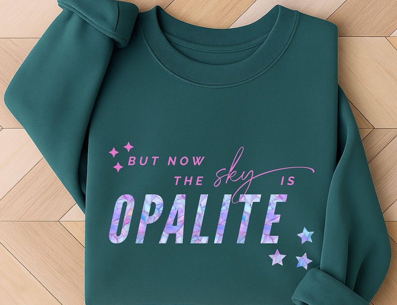 May include: Teal-colored sweatshirt with the text "But now the sky is Opalite" in a mix of fonts and colors. The word "Opalite" has a colorful, iridescent design. Pink stars and diamond shapes accent the text.