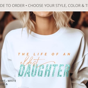 May include: White sweatshirt with the text "THE LIFE OF AN eldest DAUGHTER" in orange and teal glitter. The top text reads "MADE TO ORDER • CHOOSE YOUR STYLE, COLOR & TEXT". The sweatshirt is white and has a crew neck.