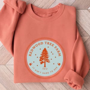 Pode incluir: Camisola coral com um gr&aacute;fico circular. O gr&aacute;fico apresenta um pinheiro laranja, rodeado pelas palavras "REDWOOD TREE FARM" e "IT AIN'T HARD TO SEE" num c&iacute;rculo azul claro.