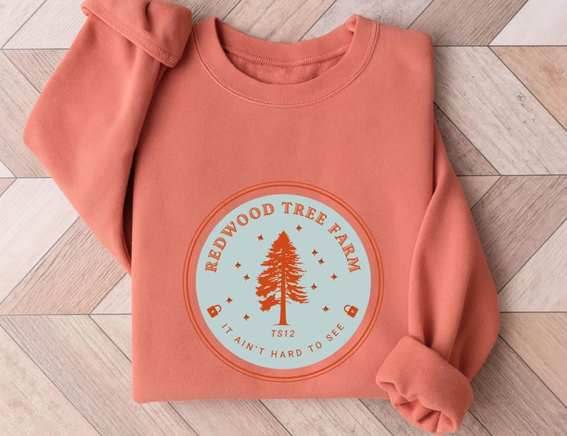 Pode incluir: Camisola coral com um gr&aacute;fico circular. O gr&aacute;fico apresenta um pinheiro laranja, rodeado pelas palavras "REDWOOD TREE FARM" e "IT AIN'T HARD TO SEE" num c&iacute;rculo azul claro.