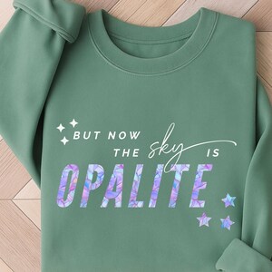 May include: Sage green sweatshirt with the text "BUT NOW THE sky IS OPALITE" in a colorful, iridescent font. The design includes star graphics. The sweatshirt has a crew neck and long sleeves.