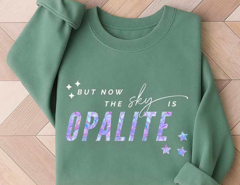 May include: Sage green sweatshirt with the text "BUT NOW THE sky IS OPALITE" in a colorful, iridescent font. The design includes star graphics. The sweatshirt has a crew neck and long sleeves.