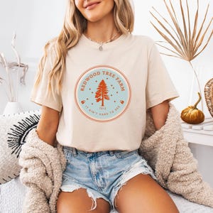Pode incluir: Camiseta bege com um gr&aacute;fico circular que diz "Redwood Tree Farm 1912 It Ain't Hard To See". O gr&aacute;fico apresenta uma &aacute;rvore vermelha dentro de um c&iacute;rculo azul claro com uma borda vermelha. A pessoa est&aacute; usando shorts jeans e um su&eacute;ter creme.