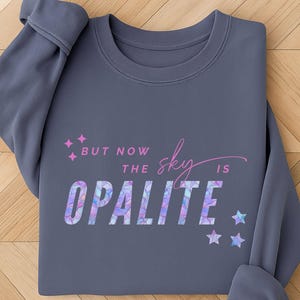 May include: A slate gray sweatshirt with the text "But now the sky is Opalite" in a colorful, geometric font. Pink star accents and a script "sky" add to the design. The sweatshirt is folded on a wooden surface.