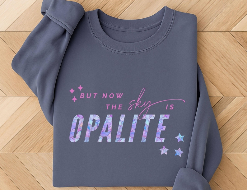 May include: A slate gray sweatshirt with the text "But now the sky is Opalite" in a colorful, geometric font. Pink star accents and a script "sky" add to the design. The sweatshirt is folded on a wooden surface.