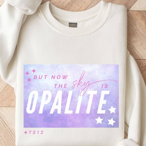 Puede incluir: Sudadera color crema con un gráfico rectangular. El gráfico presenta las palabras "BUT NOW THE sky IS OPALITE" en blanco y rosa, con un fondo de acuarela morado y azul. También hay estrellas rosas y el texto "+TS12".