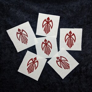 Könnte beinhalten: Sechs weiße Stoffflicken mit einem weinroten grafischen Design. Jeder quadratische Flicken zeigt ein stilisiertes Emblem mit einer zentralen Figur und flügelartigen Elementen. Die Flicken sind auf einer dunklen, strukturierten Oberfläche angeordnet.