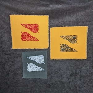 May include: Three square fabric patches with a raw edge. Two are yellow with a red design, one is gray with a white design, and one is yellow with a black design. The design on each patch is a stylized, symmetrical shape.