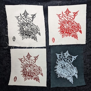 Op de afbeelding: Vier stoffen vierkanten met drakenkopontwerpen. Eén vierkant heeft een zwarte draak op een crèmekleurige achtergrond, een andere een rode draak en een derde een donkerrode draak. Het vierde vierkant heeft een witte draak op een donkergrijze achtergrond.