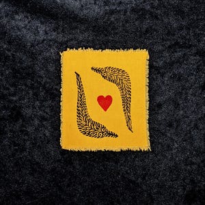 May include: A square, yellow fabric patch with a frayed edge. The patch features two black, curved, leaf-like designs framing a red heart in the center. The background is a dark, textured fabric.