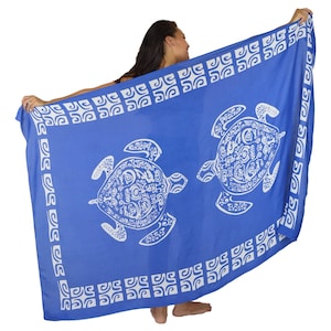 May include: A rectangular sarong in a vibrant blue colour featuring two large white sea turtles with intricate patterns on their shells. The fabric is bordered by a repeating white geometric square motif along all four edges of the textile.