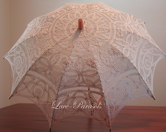 May include: A beige lace parasol featuring an intricate floral and geometric pattern throughout the canopy. The parasol has a central wooden finial at the top and the text Lace-Parasols is printed in white script across the lower front section.