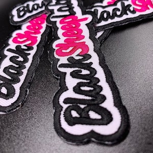 May include: Close-up of several white and black embroidered patches with the text "Black Sheep" in pink lettering.