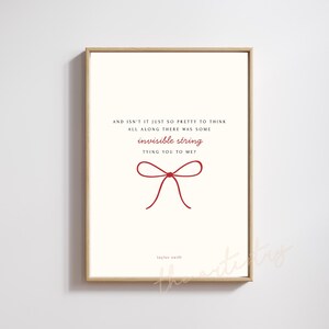 Op de afbeelding: Een ingelijste print met een crèmekleurige achtergrond toont de quote "AND ISN'T IT JUST SO PRETTY TO THINK ALL ALONG THERE WAS SOME invisible string TYING YOU TO ME?" in donkerrode tekst. Een rode strik-afbeelding is gecentreerd onder de quote, met de naam van de artiest, Taylor Swift, onderaan.