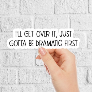 May include: A white sticker with black text that reads, "I'll get over it, just gotta be dramatic first." The sticker is held by a hand against a white brick wall background.