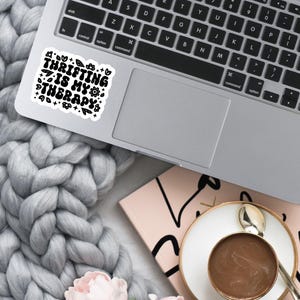 May include: A black and white sticker with the text "Thrifting is my therapy" surrounded by small floral designs. The sticker is on a silver laptop.