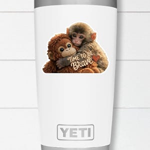 May include: White Yeti tumbler with a graphic of two hugging monkeys. The monkeys are brown and tan, with the text "TIME TO BE BRAVE" and a red heart. The tumbler has a silver rim and the Yeti logo.