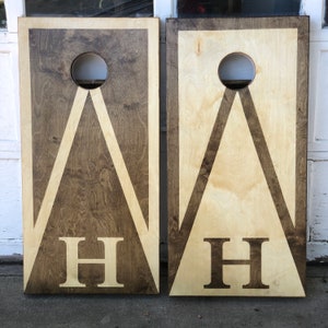May include: Two wooden cornhole boards with a white triangle design and the letter "H" in the center. The boards are stained in a dark brown color.