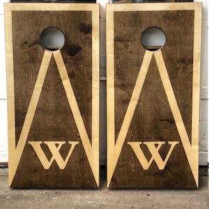 May include: Two wooden cornhole boards with a brown stain and a white triangle design. The boards have a white letter "W" in the center of each triangle.