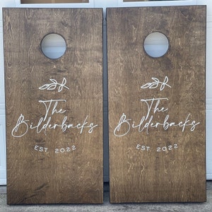 K&ouml;nnte beinhalten: Zwei braune Holz-Cornhole-Bretter mit wei&szlig;er Schrift. Der Text lautet "The Bilderbacks EST. 2022" auf jedem Brett. Jedes Brett hat ein kreisf&ouml;rmiges Loch in der Mitte.