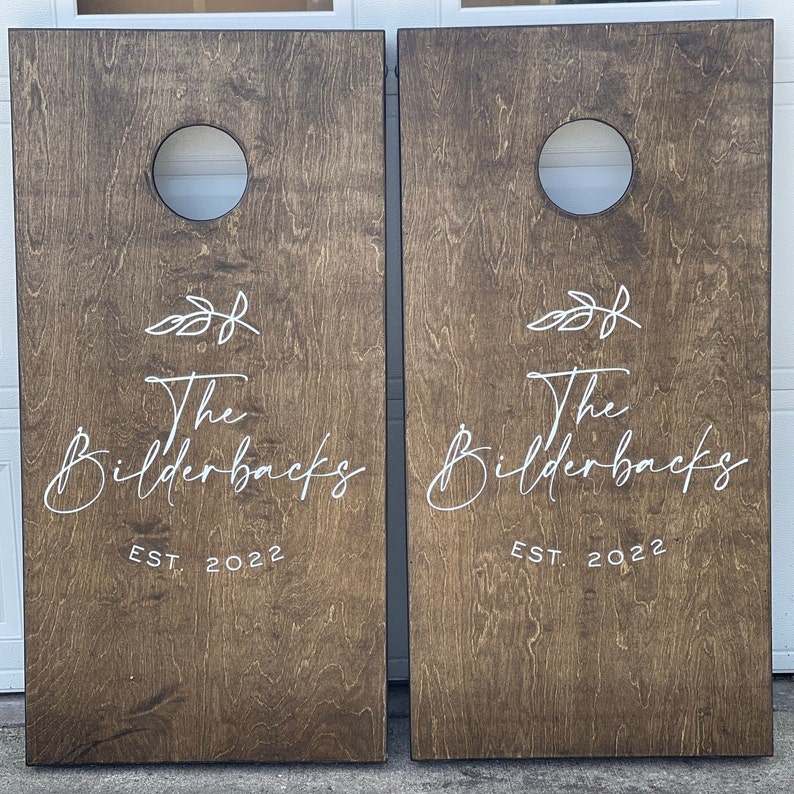 K&ouml;nnte beinhalten: Zwei braune Holz-Cornhole-Bretter mit wei&szlig;er Schrift. Der Text lautet "The Bilderbacks EST. 2022" auf jedem Brett. Jedes Brett hat ein kreisf&ouml;rmiges Loch in der Mitte.