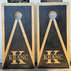 May include: Two wooden cornhole boards. Each board has a black center with a light wood border and accents. A circular hole is cut near the top. The word "KRANC" is engraved at the bottom, with a large "K" above it.