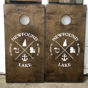 May include: Two brown wooden cornhole boards with white painted designs. The design includes the words "NEWFOUND LAKE", a small anchor, a cocktail glass, a beach chair, and the coordinates "43.0469° N 71.8056° W".