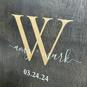 May include: A dark wood sign with a large white "W" in the center. The text "amy + mark" is written in white cursive script below the "W". The date "03.24.24" is written in white below the text.