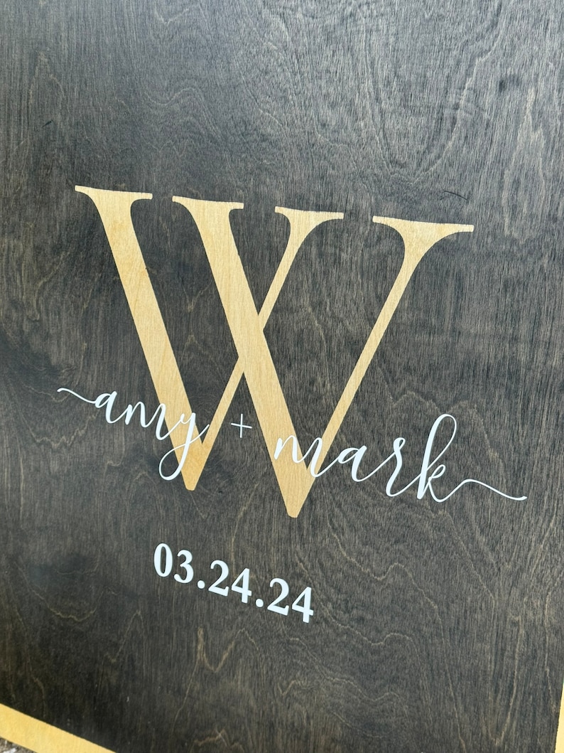 May include: A dark wood sign with a large white "W" in the center. The text "amy + mark" is written in white cursive script below the "W". The date "03.24.24" is written in white below the text.