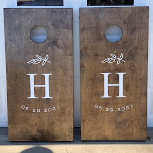 May include: Two brown wooden cornhole boards with white painted designs. Each board has a white circle in the center, a white leafy branch design, the letter "H" and the date "05.29.2021".