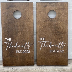 May include: Two brown wooden cornhole boards with white painted text that reads "THE Thibaults EST. 2022".