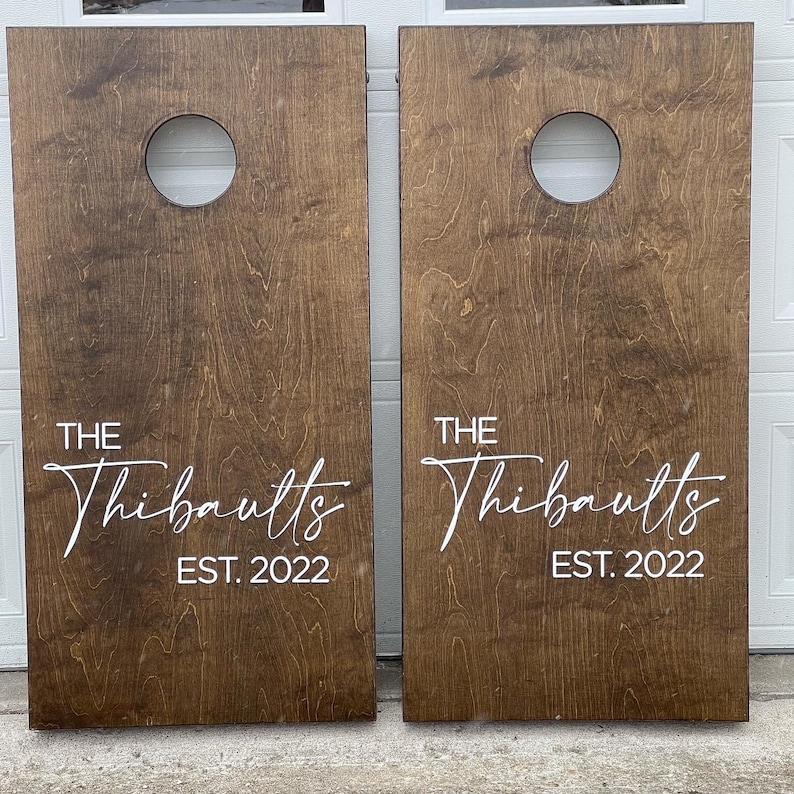 May include: Two brown wooden cornhole boards with white painted text that reads "THE Thibaults EST. 2022".