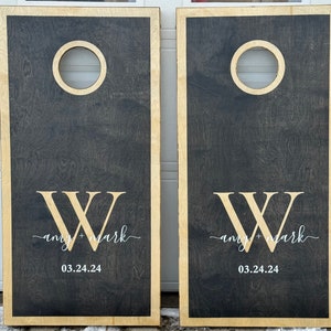 May include: Two black wooden cornhole boards with a white painted "W" and the text "any + mark" and "03.24.24" on each board.
