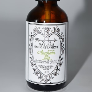 Könnte beinhalten: Eine braune Glasflasche mit schwarzem Sprühkopf und einem weißen Etikett. Das Etikett hat ein herzförmiges Design mit einem Pfeil und dem Text "NATURE'S ENLIGHTENMENT Anghalata Lily Green Aventurine & Tibetan Clear Quartz HEART CHAKRA GEM ELIXIR SPRAY 2 oz".