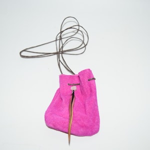 K&ouml;nnte beinhalten: Ein kleiner rosa Wildlederbeutel mit einem braunen Lederzugband und einem Federschmuck. Der Beutel h&auml;ngt an einem braunen Lederband.