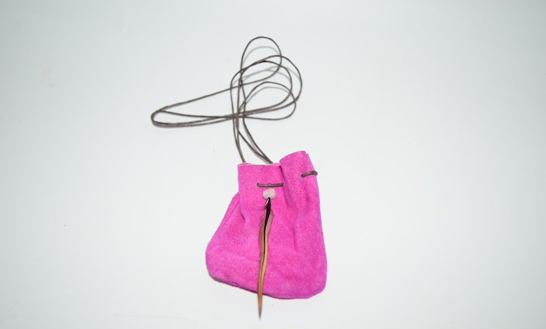 K&ouml;nnte beinhalten: Ein kleiner rosa Wildlederbeutel mit einem braunen Lederzugband und einem Federschmuck. Der Beutel h&auml;ngt an einem braunen Lederband.
