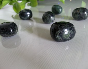 May include: A collection of six smooth, dark green and black tumbled stones with a speckled pattern. The stones are arranged on a white surface.