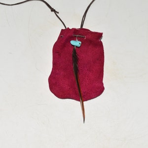 K&ouml;nnte beinhalten: Ein kleiner roter Lederbeutel mit einer braunen Lederschnur und einer t&uuml;rkisfarbenen Perle. Eine einzelne braune Feder h&auml;ngt an dem Beutel.