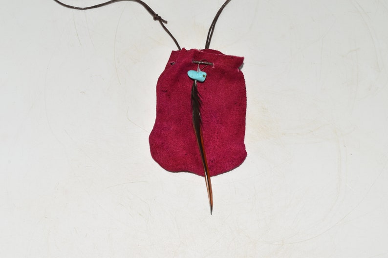 K&ouml;nnte beinhalten: Ein kleiner roter Lederbeutel mit einer braunen Lederschnur und einer t&uuml;rkisfarbenen Perle. Eine einzelne braune Feder h&auml;ngt an dem Beutel.